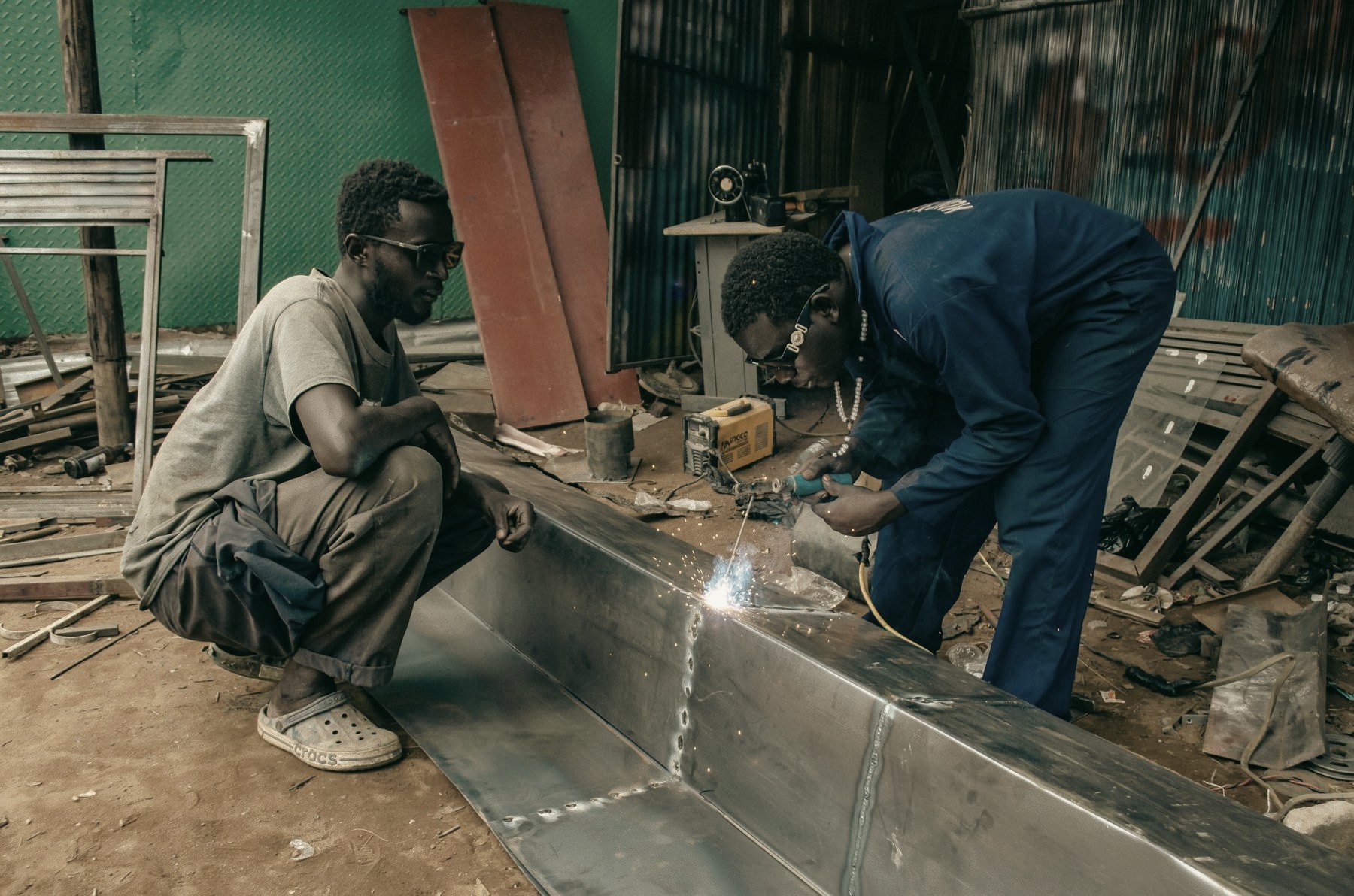 Two men in a metal workshop, one inspecting the work while the other welds